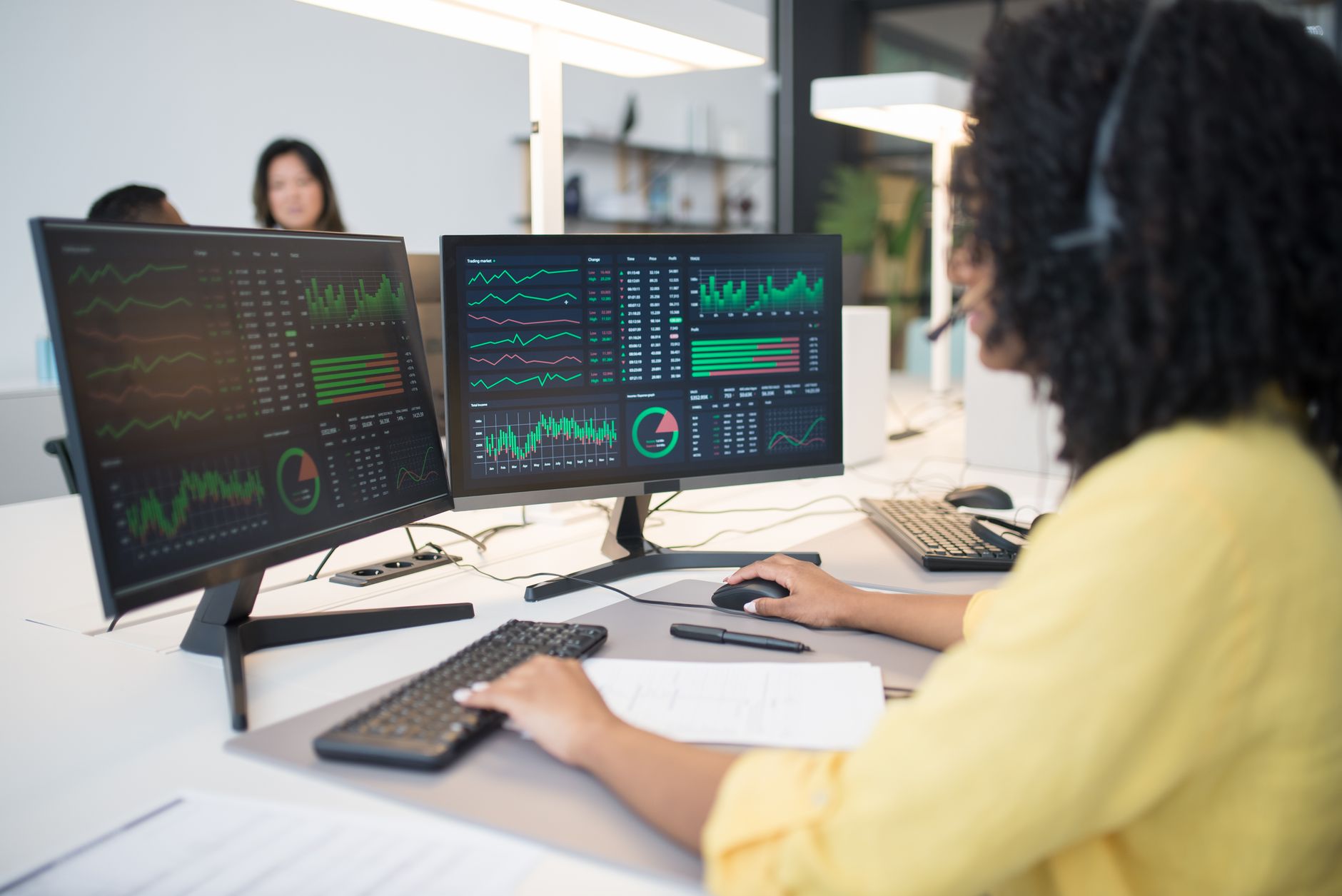 An operations analyst at her desk with two monitoring screens, reading the day's figures